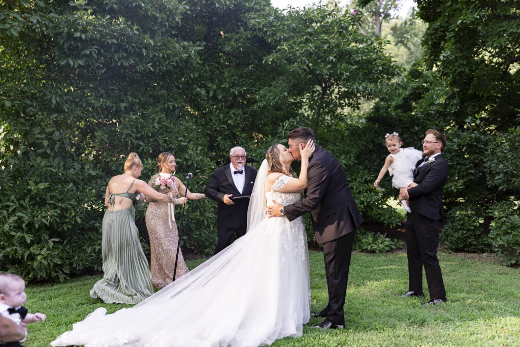 The newlyweds share their first kiss on a grassy ceremony lawn surrounded by dense greenery, while members of the wedding party look on and a toddler is held nearby during their Pennsylvania garden wedding.