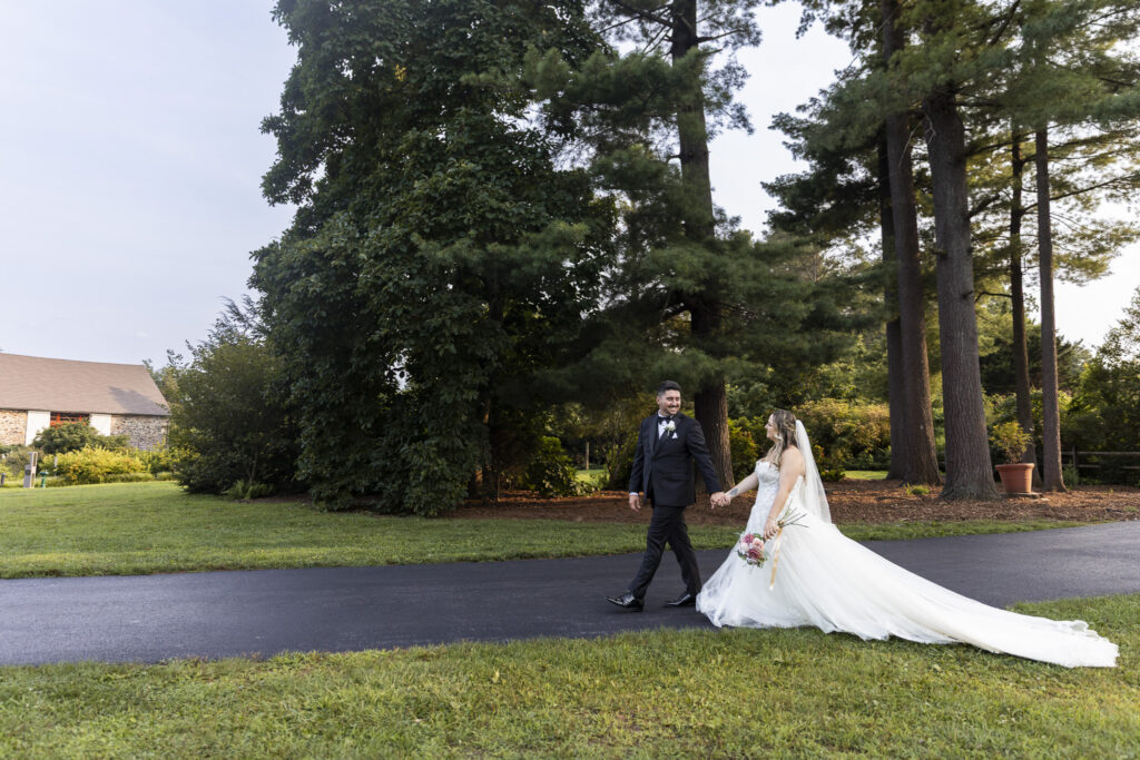 Bride and groom walk hand in hand through the expansive green grounds after their Tyler Arboretum wedding, with tall trees and a stone barn in the background.