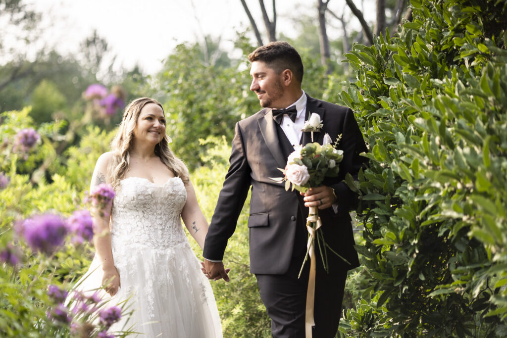The newlyweds walk hand in hand along a narrow garden path surrounded by tall greenery and purple flowers, smiling at one another while the groom carries a pink and white bouquet during their Pennsylvania garden wedding.