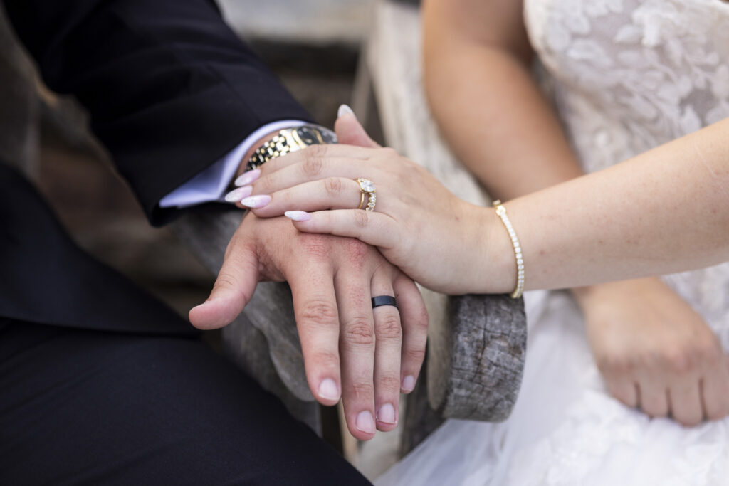 Close-up of the newlyweds’ hands resting together on a wooden bench, highlighting their wedding rings. The bride’s oval engagement ring and diamond wedding band are visible alongside her pale pink manicure and delicate bracelet, while the groom wears a dark wedding band and metal wristwatch.