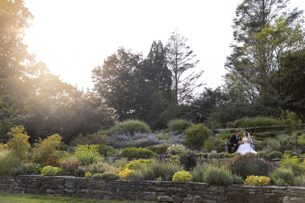 Wide portrait of the bride and groom sitting together in the lush terraced gardens during golden hour at their Tyler Arboretum wedding, surrounded by colorful flowers, stone walls and mature trees as the sun shines directly at the camera on the opposite side