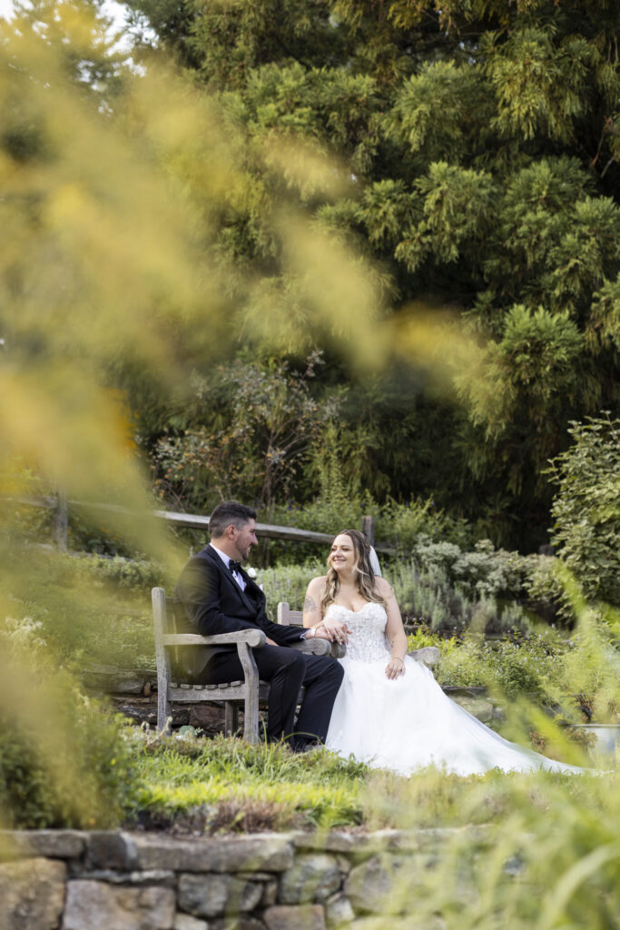 Bride and groom sit together on a wooden garden bench surrounded by layered flowering plants and stone walls in the gardens at Tyler Arboretum.