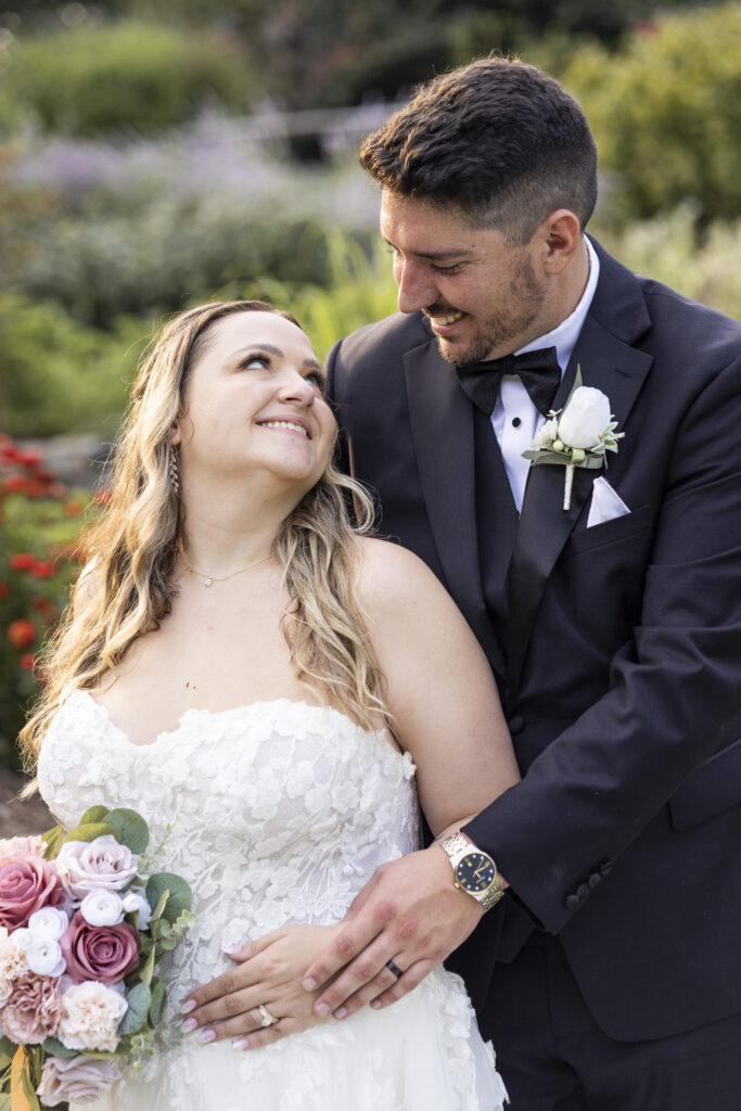 The bride and groom smile warmly at one another during portraits in the gardens, her pink and mauve bouquet visible beside her lace gown and his white rose boutonniere complementing his black tuxedo at their Pennsylvania garden wedding.