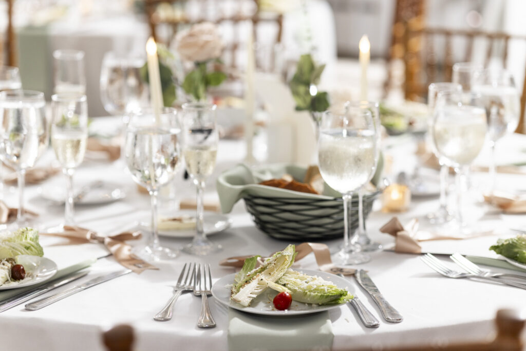 A sunlit wedding reception table is set with white linens, sage green napkins, blush ribbon details, glassware, silverware, candles, and plates of salad. A dark woven bread basket sits near the center among soft floral arrangements and glowing taper candles.