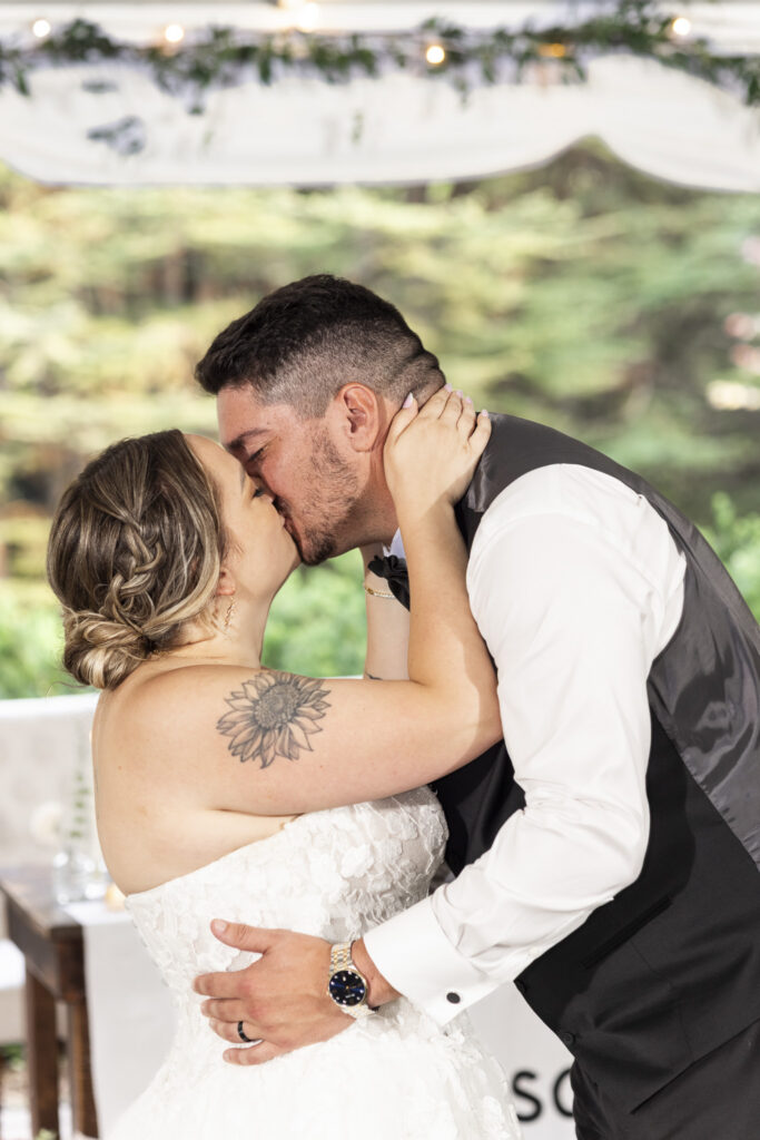 Close-up of the newlyweds kissing during their first dance, with the bride’s floral shoulder tattoo, braided updo and lace wedding gown visible beneath the reception tent at their Pennsylvania garden wedding.