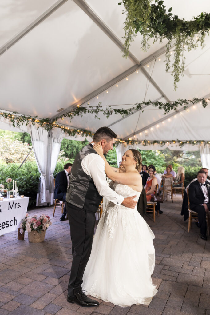 The bride and groom hold one another during their first dance beneath a white reception tent decorated with hanging greenery, string lights and open views of the surrounding gardens at their Pennsylvania garden wedding.