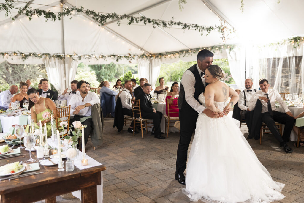 The groom wraps his arms around the bride and kisses the top of her head during their wedding reception beneath a white tent. Guests sit around decorated dinner tables as the newlyweds share a quiet affectionate moment surrounded by family and friends.