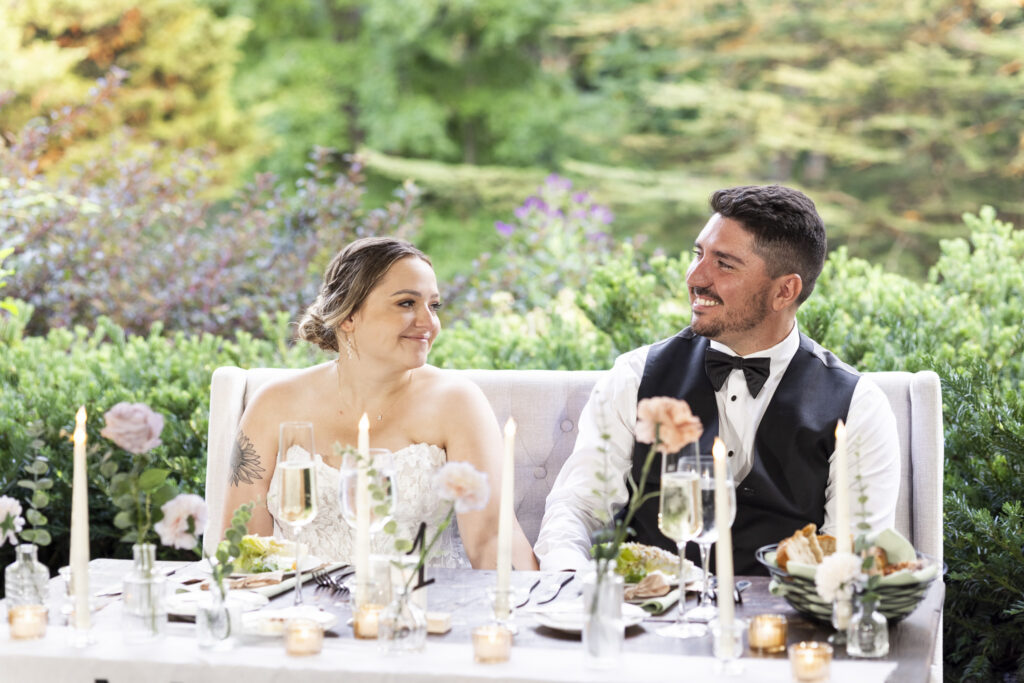 The bride looks at her groom and smiles at their reception table, surrounded by taper candles, glassware and soft floral arrangements overlooking the greenery of their Pennsylvania garden wedding.