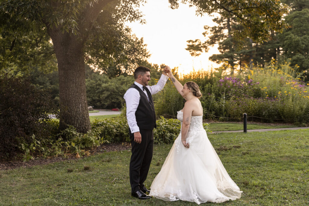 The newlyweds share a playful golden hour wedding portrait in the gardens as the groom raises the bride’s hand and twirls her beneath it. Her full lace wedding gown spreads across the grass while warm sunset light filters through the trees and illuminates the flowering garden behind them.