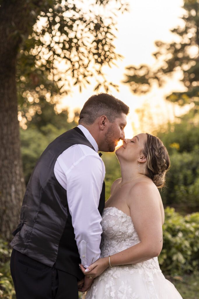 The newlyweds lean toward one another for a kiss during golden hour wedding portraits in the garden. Warm sunlight shines between their faces and through the surrounding trees as they hold hands, with the bride’s lace gown and braided updo softly illuminated by the setting sun.
