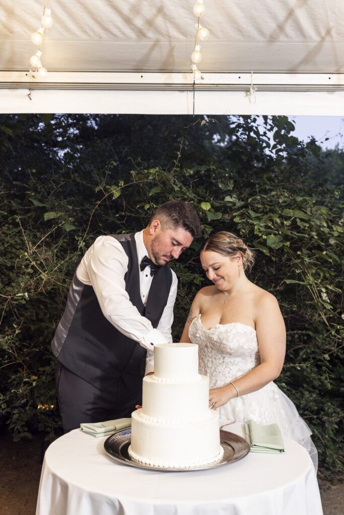 The newlyweds stand together behind a simple three-tier white wedding cake, guiding the knife through the bottom tier beneath string lights with dense summer greenery behind them at their Pennsylvania garden wedding.