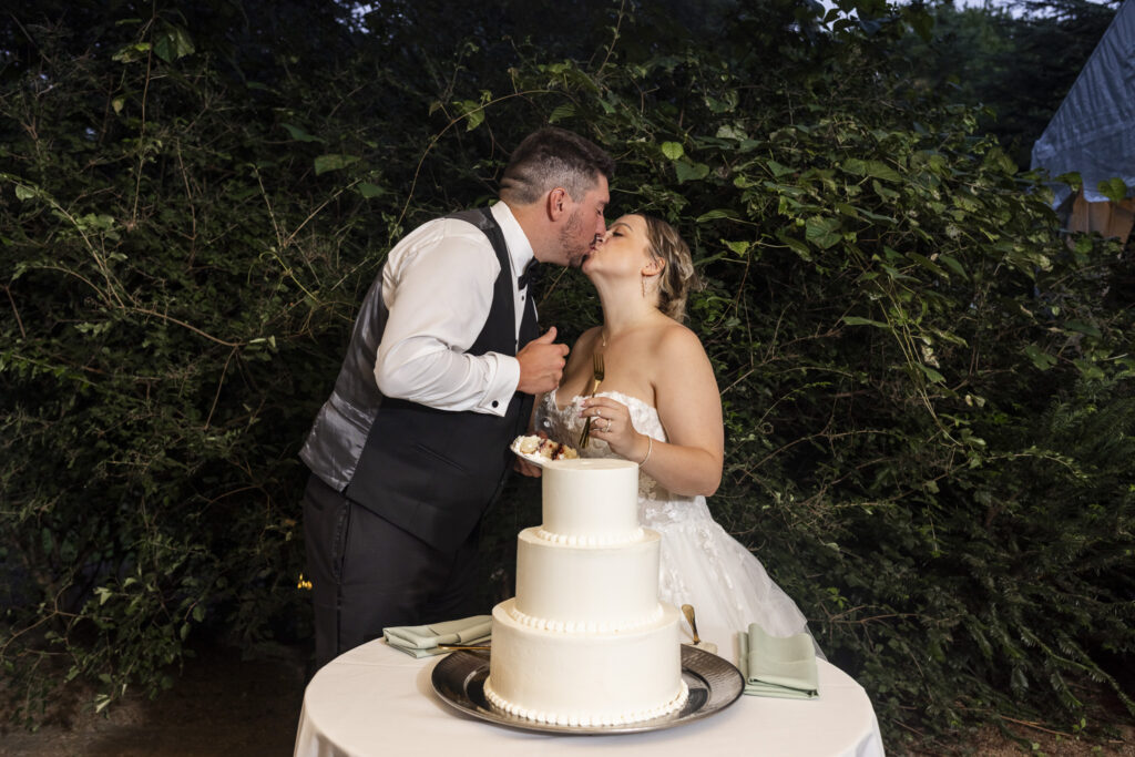 The newlyweds lean across their three-tier wedding cake to kiss after cutting a slice, surrounded by dark leafy greenery during the evening reception at their Pennsylvania garden wedding.
