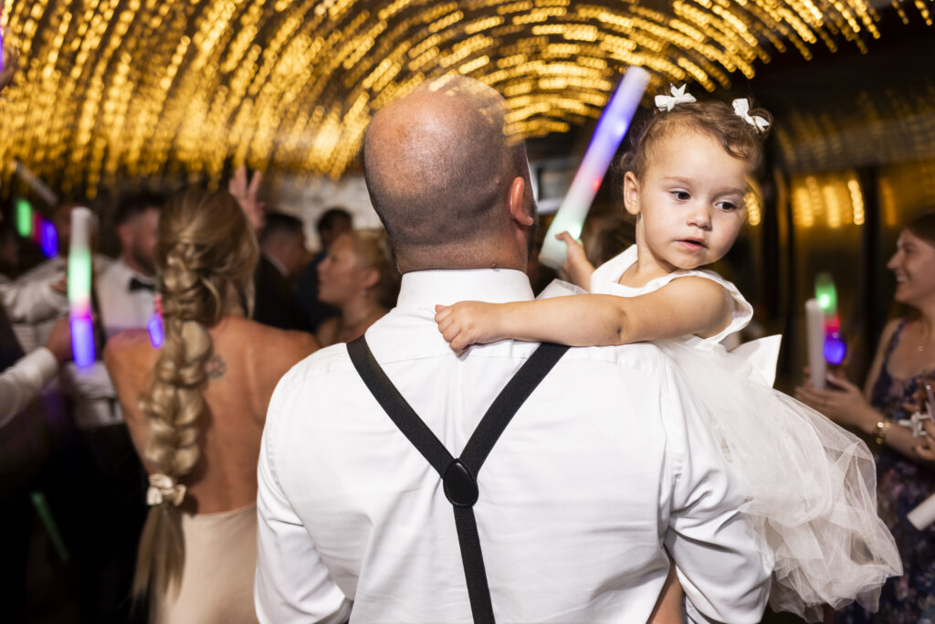 A toddler in a white tulle dress is carried across the crowded dance floor as colorful glow sticks and swirling golden lights fill the reception during a Pennsylvania garden wedding.