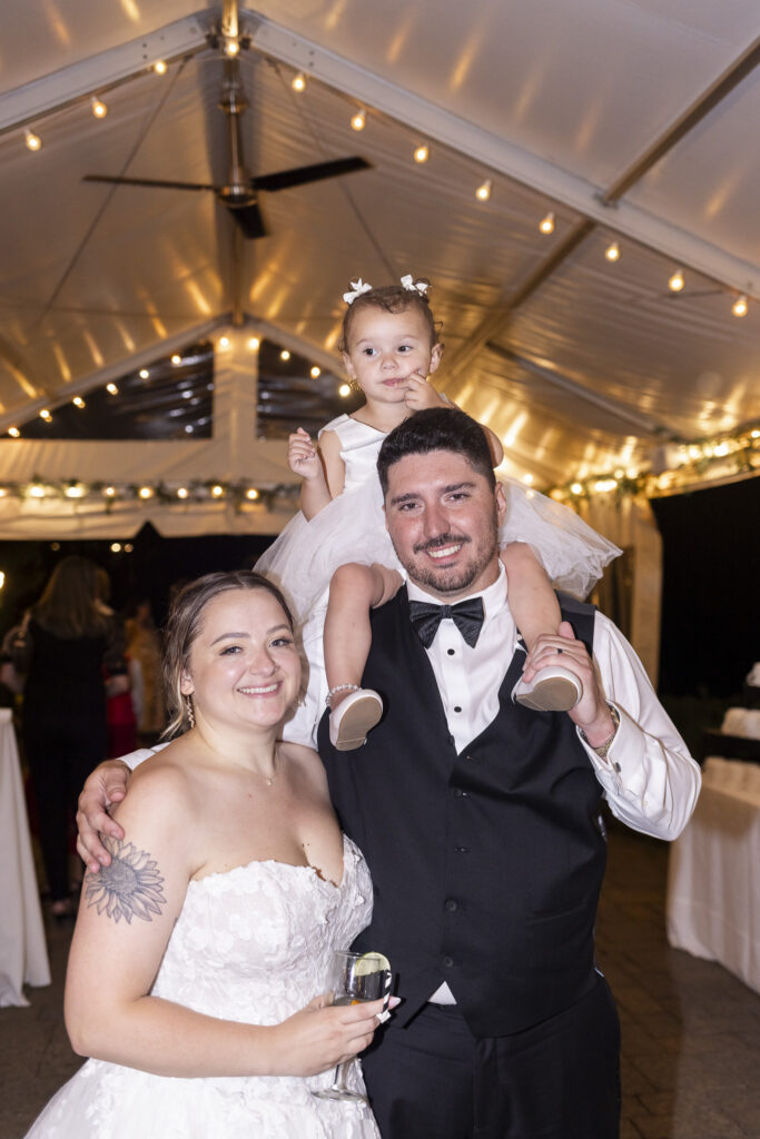 The bride and groom smile beneath warm string lights at their evening wedding reception while a young child in a white dress sits on the groom’s shoulders. The bride holds a drink beside them, creating a playful family moment during the celebration.