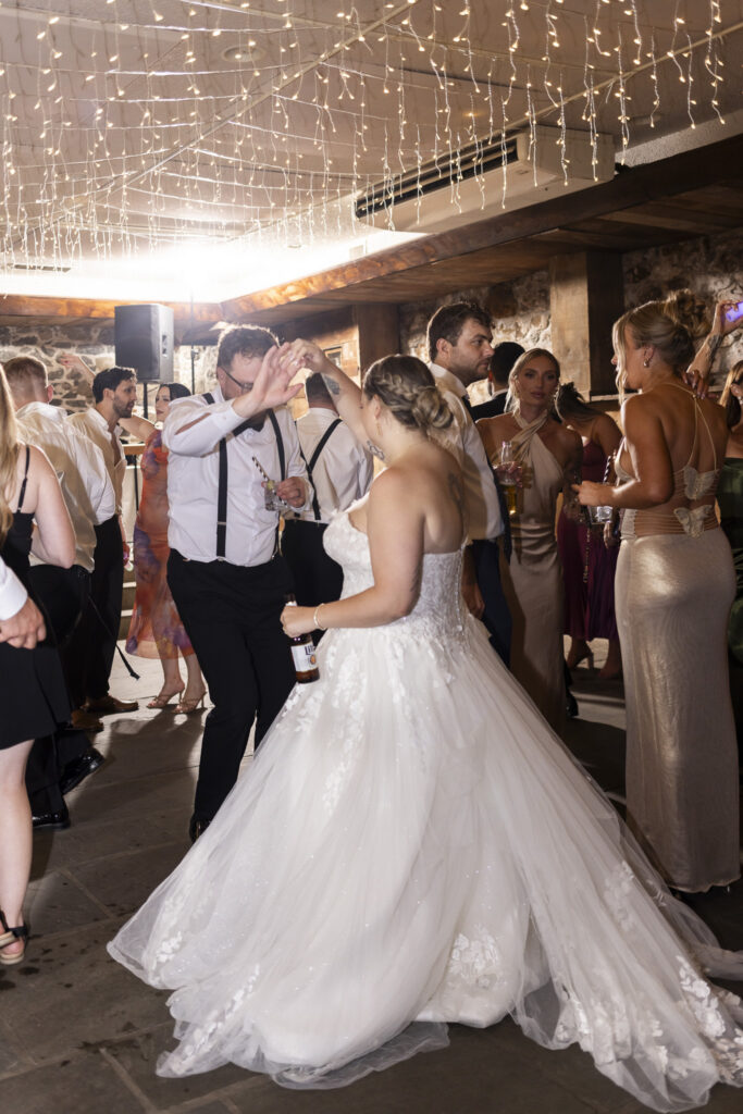 The bride dances with a guest beneath strands of warm white lights as friends gather around the lively stone-walled reception dance floor following their Pennsylvania garden wedding.