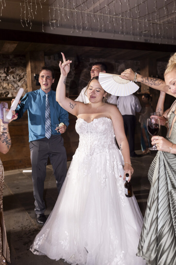 The bride dances with one arm raised during the wedding reception while holding a beer bottle in her other hand. Friends surround her on the dance floor, including a guest holding an open white folding fan above her as the group celebrates together.