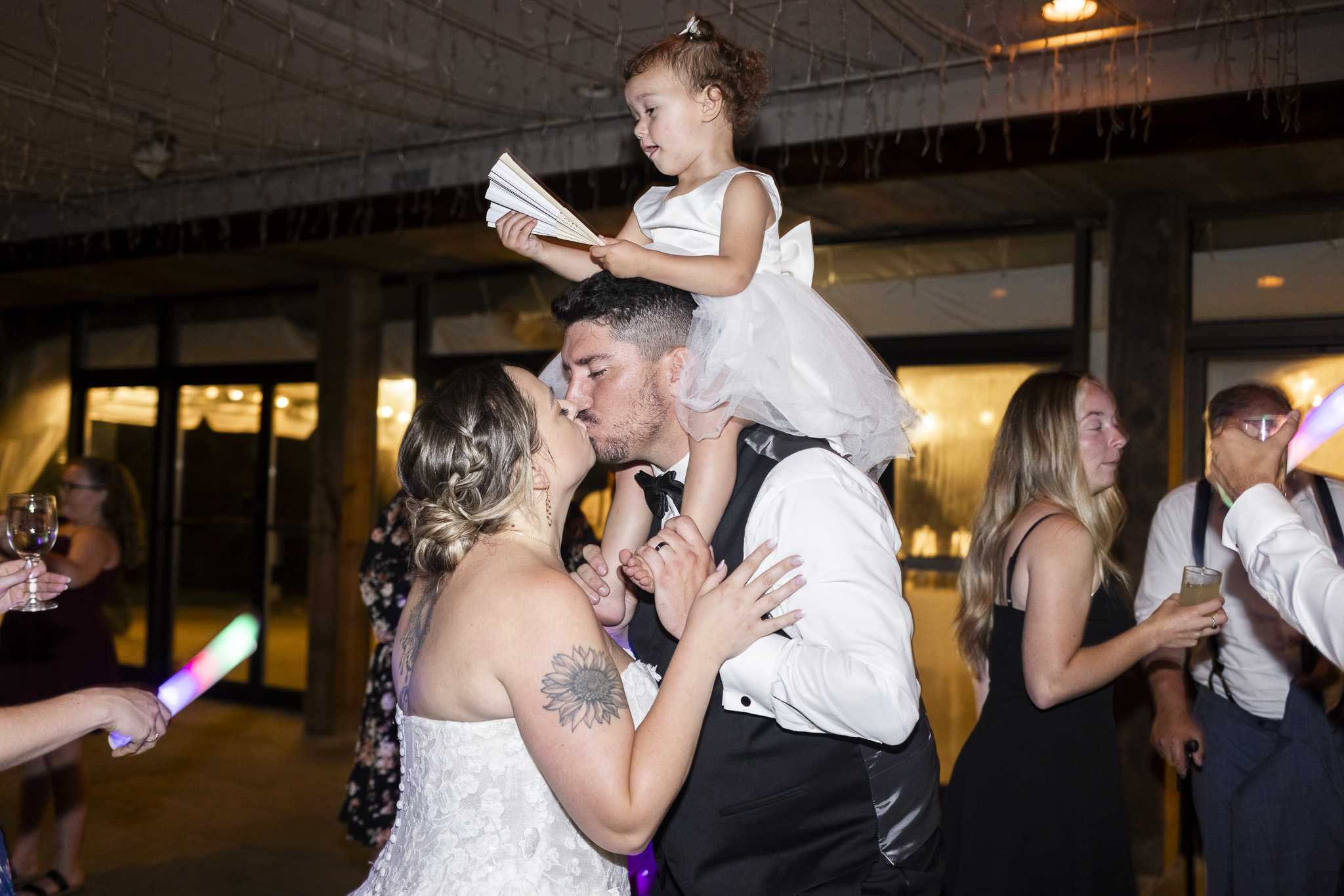 Bride and groom kiss on the reception dance floor while their toddler sits on the groom’s shoulders holding an open folding fan.