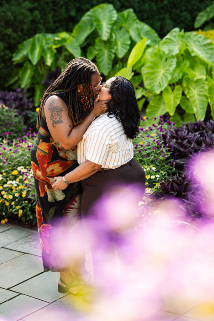 Newly engaged couple sharing a kiss after their Longwood Gardens proposal, surrounded by oversized green leaves and small purple and yellow flowers, with soft pink blooms blurred across the foreground.