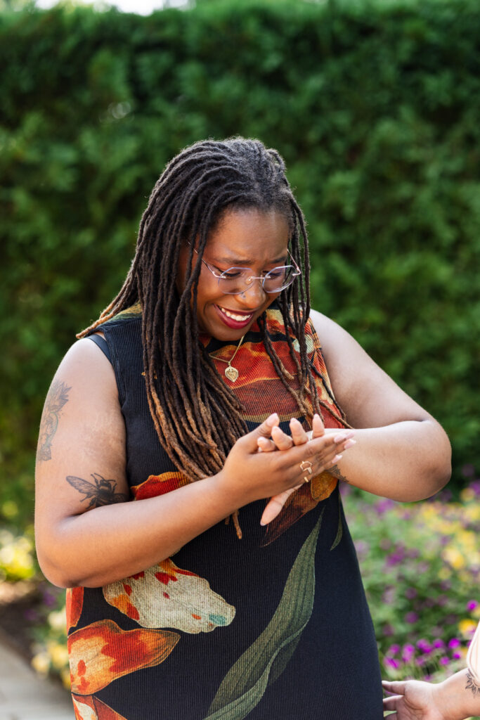 Emotional reaction during a Longwood Gardens lesbian proposal as the newly engaged partner looks down at the ring on her hand, smiling through tears against a backdrop of hedges and colorful flowers.