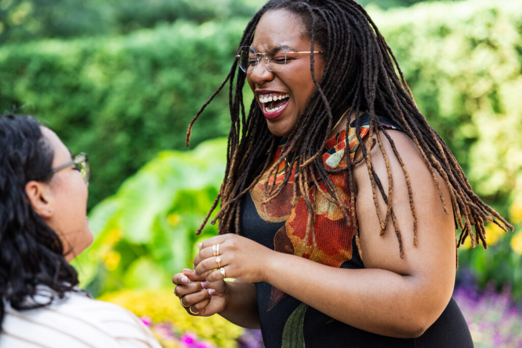 Joyful moment after a lesbian proposal, with the newly engaged partner laughing broadly and holding up her ring hand as her fiancée smiles toward her among bright garden foliage.