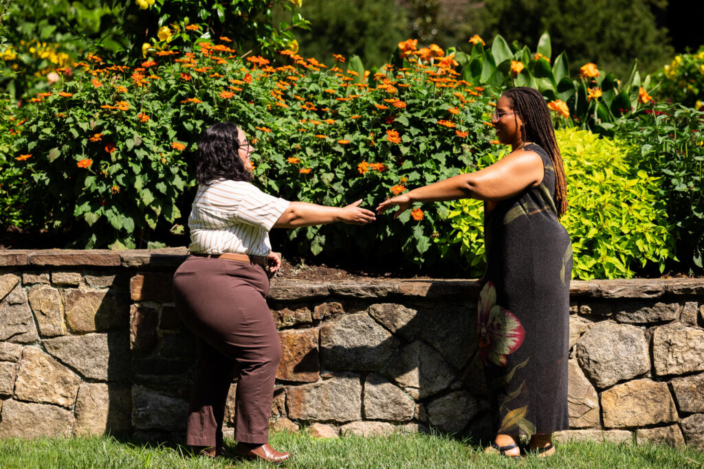 Playful Longwood Gardens lesbian proposal portrait with the couple reaching toward one another in front of a stone garden wall, backed by masses of vivid orange flowers and chartreuse foliage.