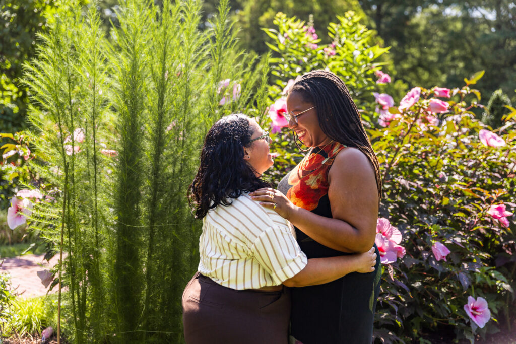 Engaged couple embracing and laughing among sunlit pink flowers during their Longwood Gardens lesbian proposal, with tall feathery greenery and glowing summer foliage surrounding them.