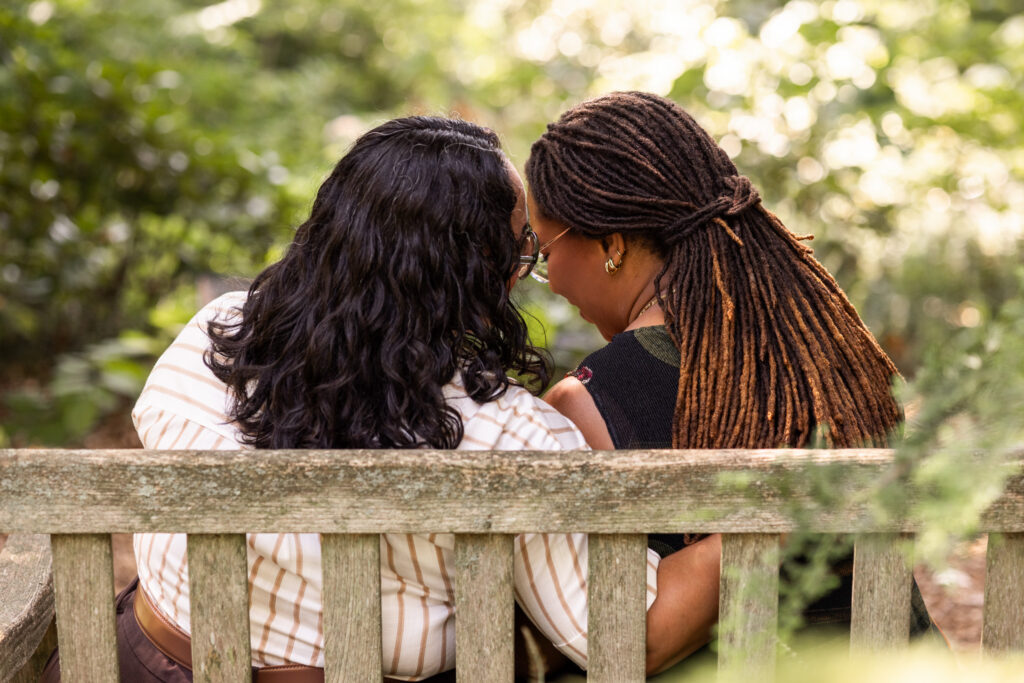 couple sitting closely together on a weathered wooden garden bench, photographed from behind as they lean their heads together beneath softly lit trees.
