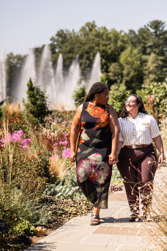 Engaged couple walking hand in hand through colorful flower beds during their Longwood Gardens lesbian proposal, smiling at each other with tall fountains spraying in the distance.