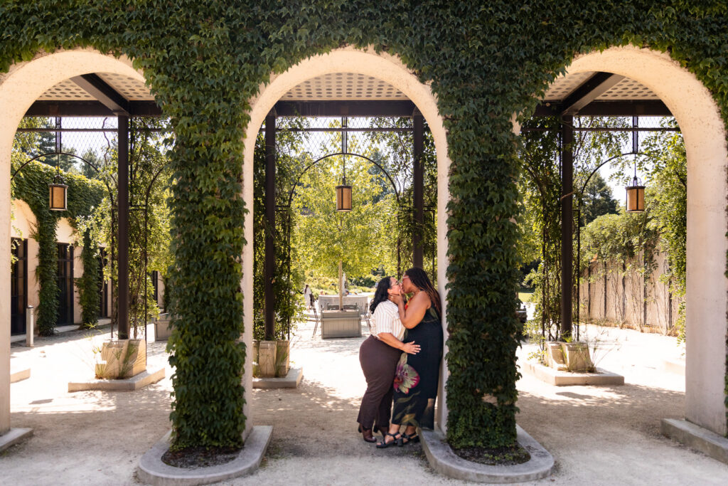 Wide lesbian proposal photography portrait of the engaged couple kissing beneath ivy-covered arches and hanging lanterns, surrounded by the formal garden architecture at Longwood Gardens.