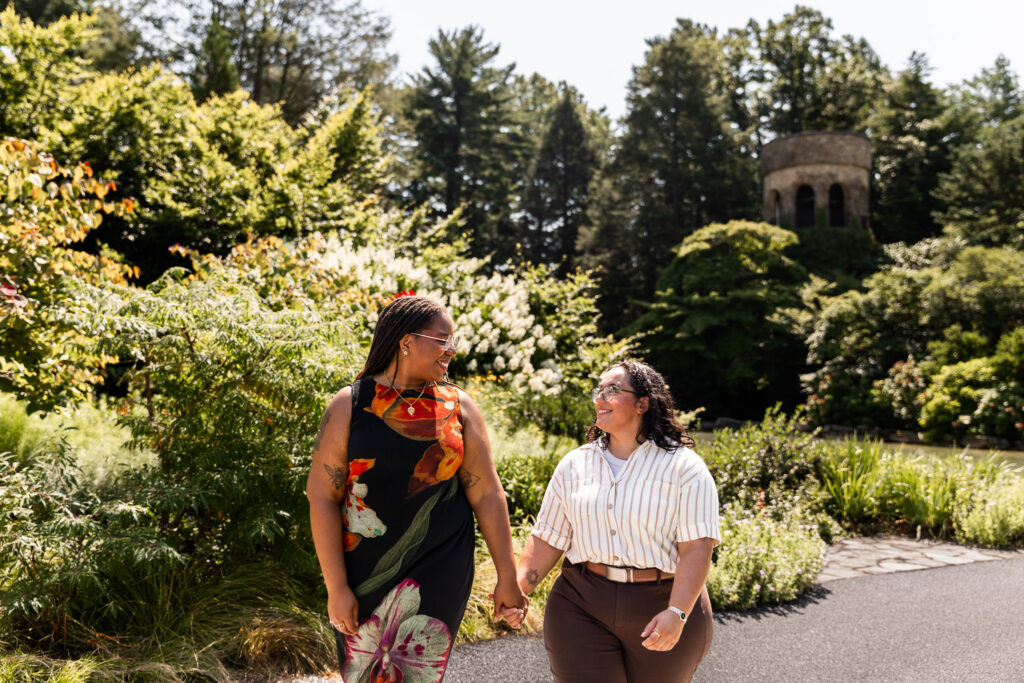 Couple walking hand in hand through lush summer gardens after their Longwood Gardens lesbian proposal, smiling at one another with dense plantings and a round stone tower rising among the trees behind them.