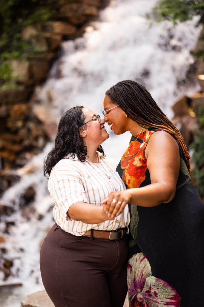 Newly engaged couple nuzzle noses while showing off the ring during their Longwood Gardens lesbian proposal portraits, holding hands in front of a softly blurred waterfall surrounded by rock and greenery.