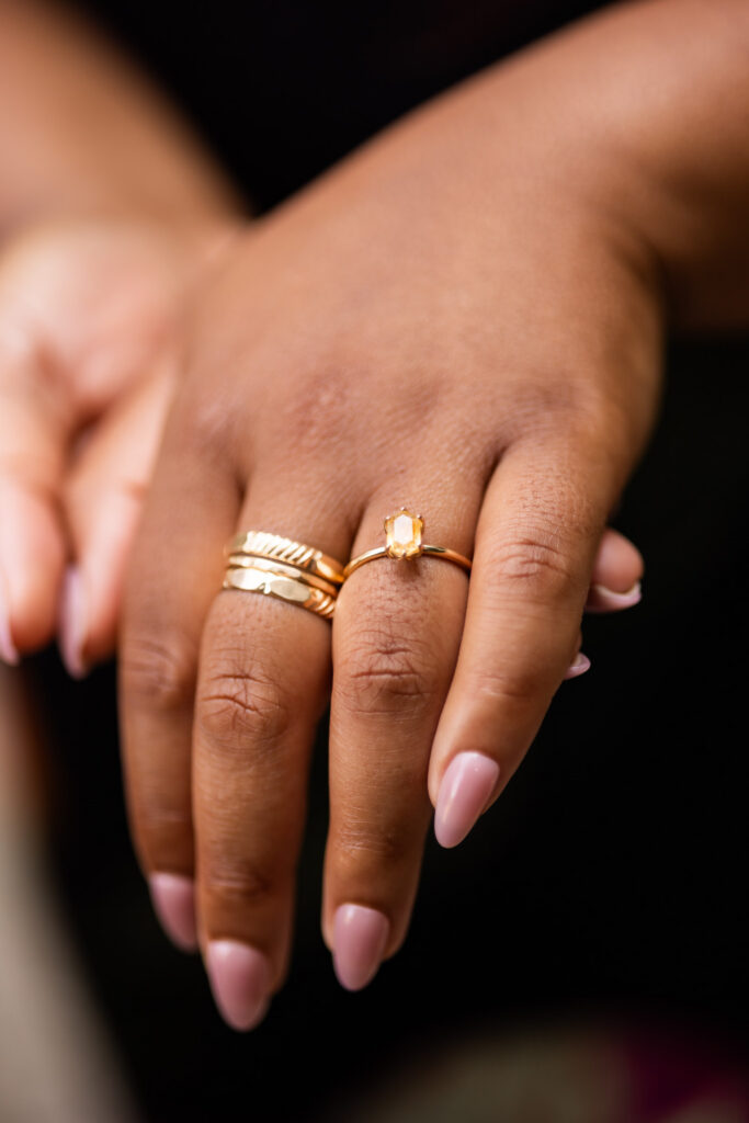Close-up showing a warm golden rectangular engagement stone on a slender gold band, paired with stacked gold rings and softly manicured pink nails.