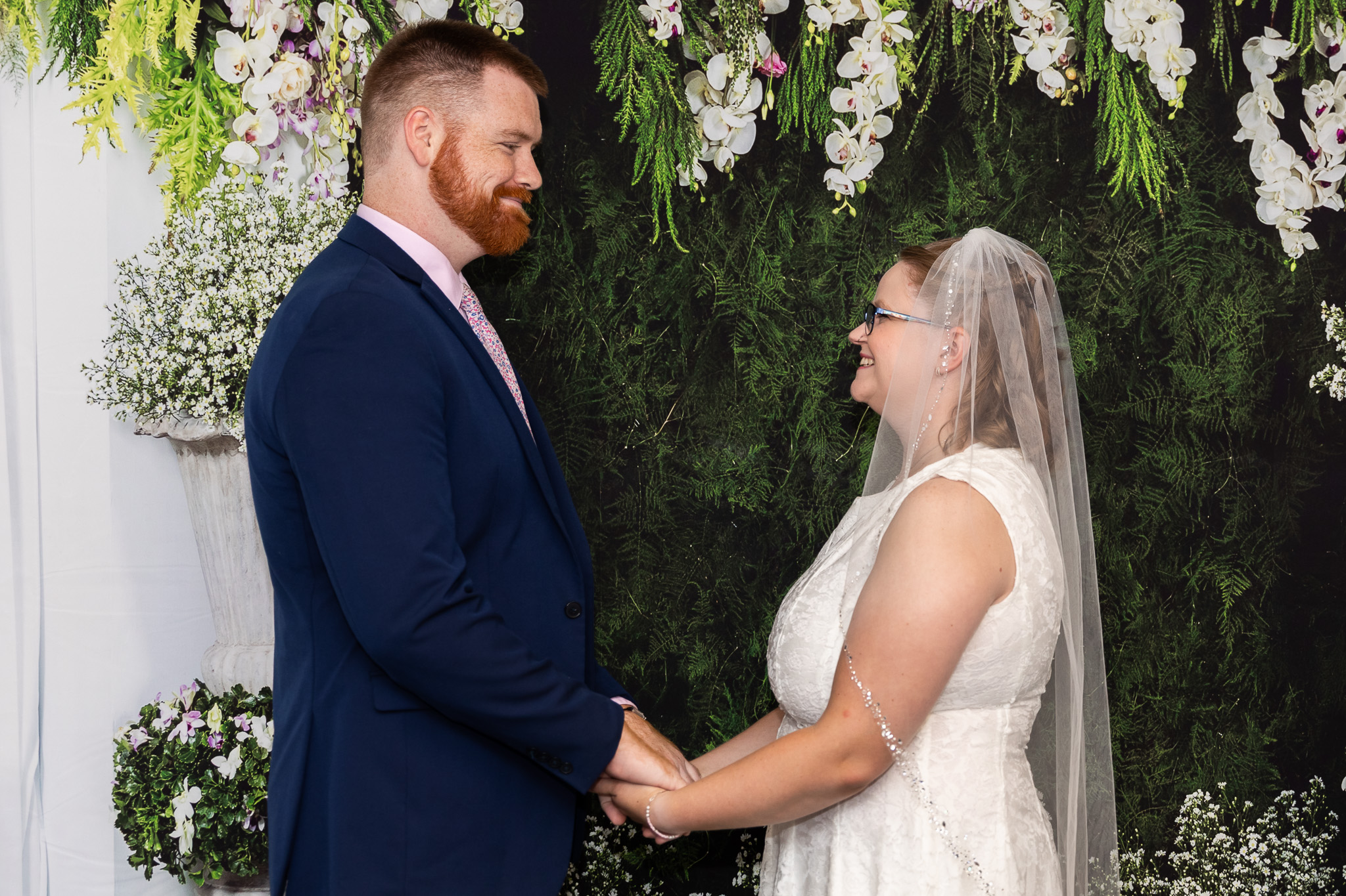 Couple holds hands and smiles at each other during their civil ceremony, with courthouse wedding photography documenting the moment beneath cascading white and purple flowers and greenery.