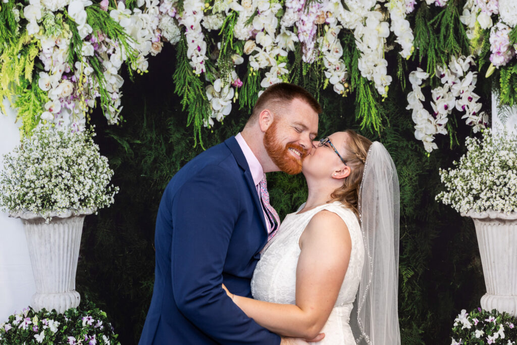 Bride in a short white lace dress and veil kisses the groom’s cheek during courthouse wedding photography, framed by a lush backdrop of white flowers and greenery.