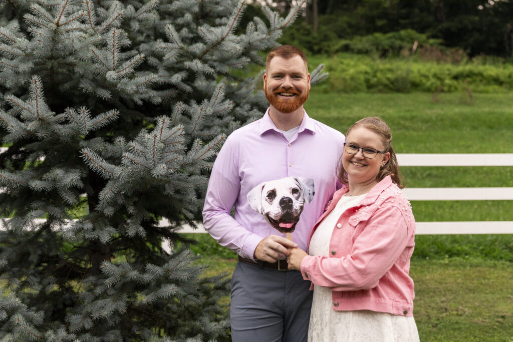 Couple smiles together during their engagement photos at Pleasant View Farm while holding a cutout of their dog’s face, standing beside a blue spruce tree and white farm fence.