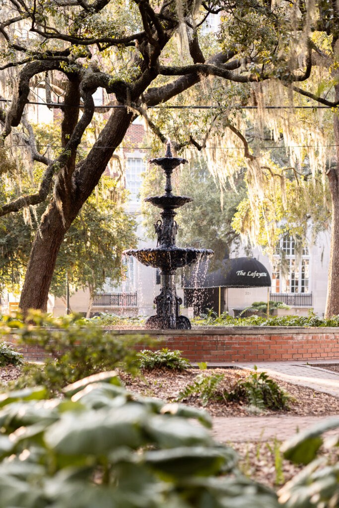 Warm golden-hour light filters through sprawling live oak branches and hanging Spanish moss around the ornate black fountain in Lafayette Square, scouted by a Savannah proposal photographer ahead of the surprise.
