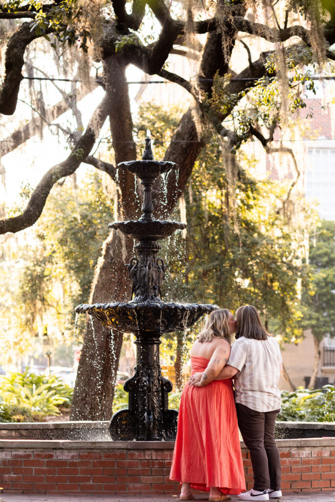 Two women stand beside the black fountain at Lafayette Square and kiss with their arms wrapped around each other. One wears a flowing coral strapless dress and the other a light striped button-up shirt and dark pants, while golden sunlight glows through the oak trees behind them just before their LGBTQ proposal in Savannah.