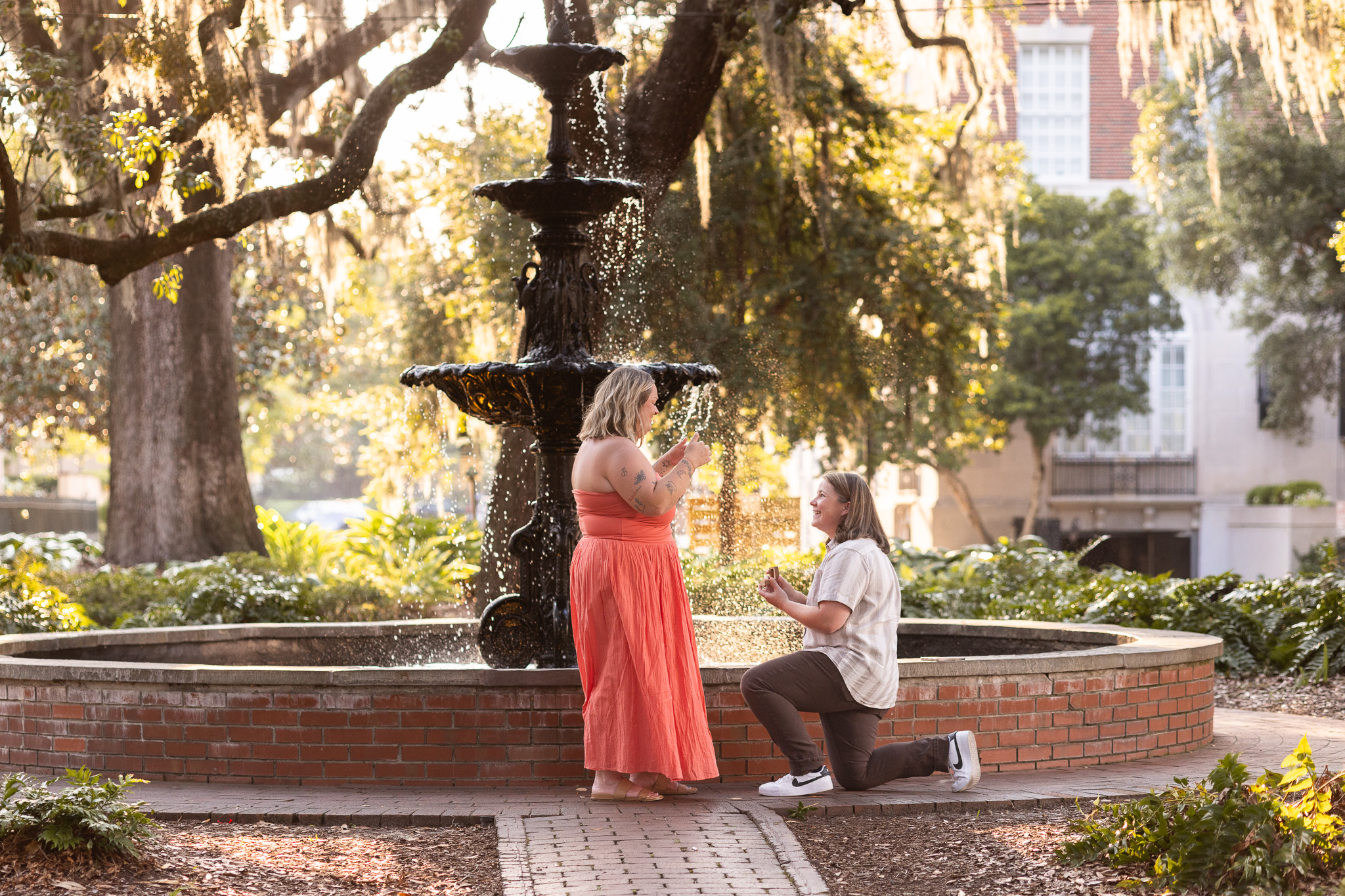 An LGBTQ proposal in Savannah unfolds beside the Lafayette Square fountain as one woman kneels with an open ring box and smiles up at her partner, who stands in a coral dress with her hands raised in surprise.