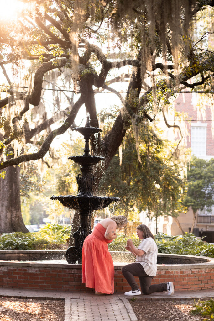Golden sunlight streams through the oak trees during an LGBTQ proposal in Savannah, where one woman bends toward her kneeling partner and covers her mouth as she realizes she is being proposed to beside the fountain.