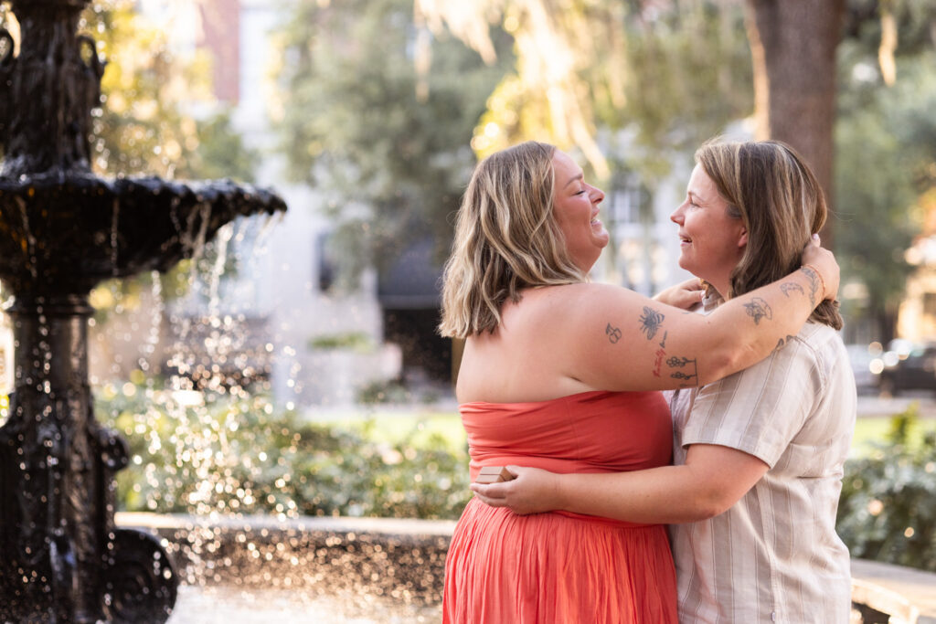 Moments after their LGBTQ proposal in Savannah, the newly engaged couple embraces beside the Lafayette Square fountain, smiling at each other while one still holds the small ring box behind her partner’s waist.