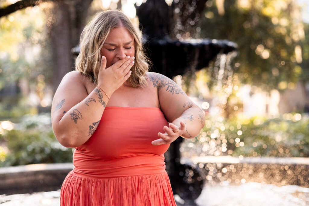 A newly engaged woman in a coral strapless dress covers her mouth with one hand while extending the other to stare at her new engagement ring, her emotional expression in sharp focus against the softly blurred fountain.