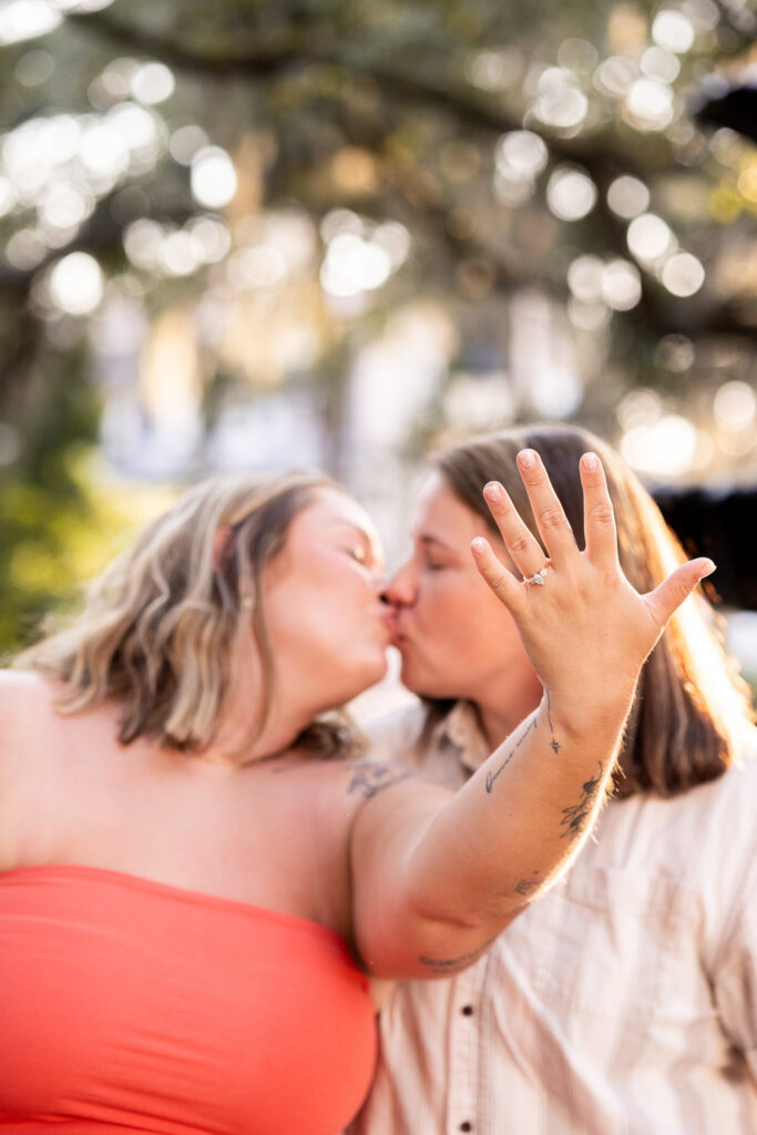 A close portrait after an LGBTQ proposal in Savannah focuses on a newly engaged woman’s raised hand and sparkling ring while she kisses her partner in the softly blurred golden-hour background.