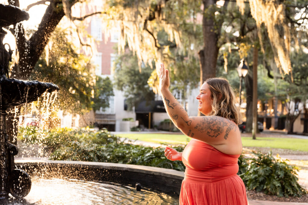 A newly engaged woman stands beside the fountain with a wide smile and raises her left hand high to excitedly show off her new ring, with warm sunlight illuminating the falling water and Spanish moss behind her.