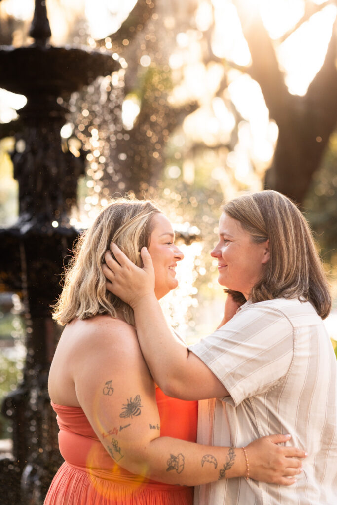 Warm backlight surrounds two women after their LGBTQ proposal in Savannah as they hold each other beside the Lafayette Square fountain, one gently cupping her partner’s face while they smile at each other.