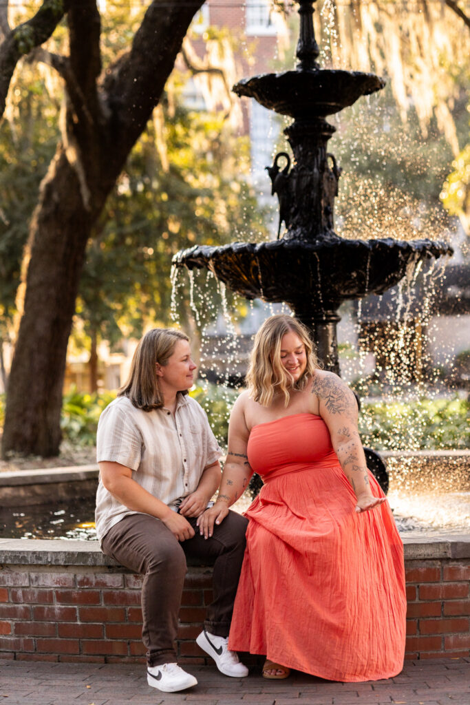 Two newly engaged women sit together on the brick edge of the Lafayette Square fountain, one in a light striped button-up shirt and brown pants looking warmly toward her partner, who wears a flowing coral strapless dress and looks down at her newly placed engagement ring. Sunlit water sparkles behind them as it falls from the ornate black fountain, framed by live oak trees and softly glowing Spanish moss.