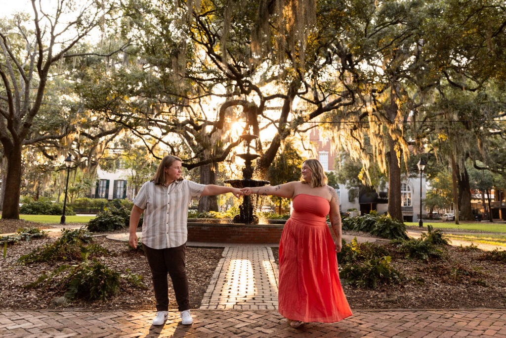 Newly engaged couple stands several feet apart on a brick walkway in Lafayette Square, reaching toward each other to hold hands with their arms fully extended. The low golden sun shines through sprawling live oak branches and hanging Spanish moss behind them, silhouetting the fountain at the center of the square and casting long shadows across the brick path.