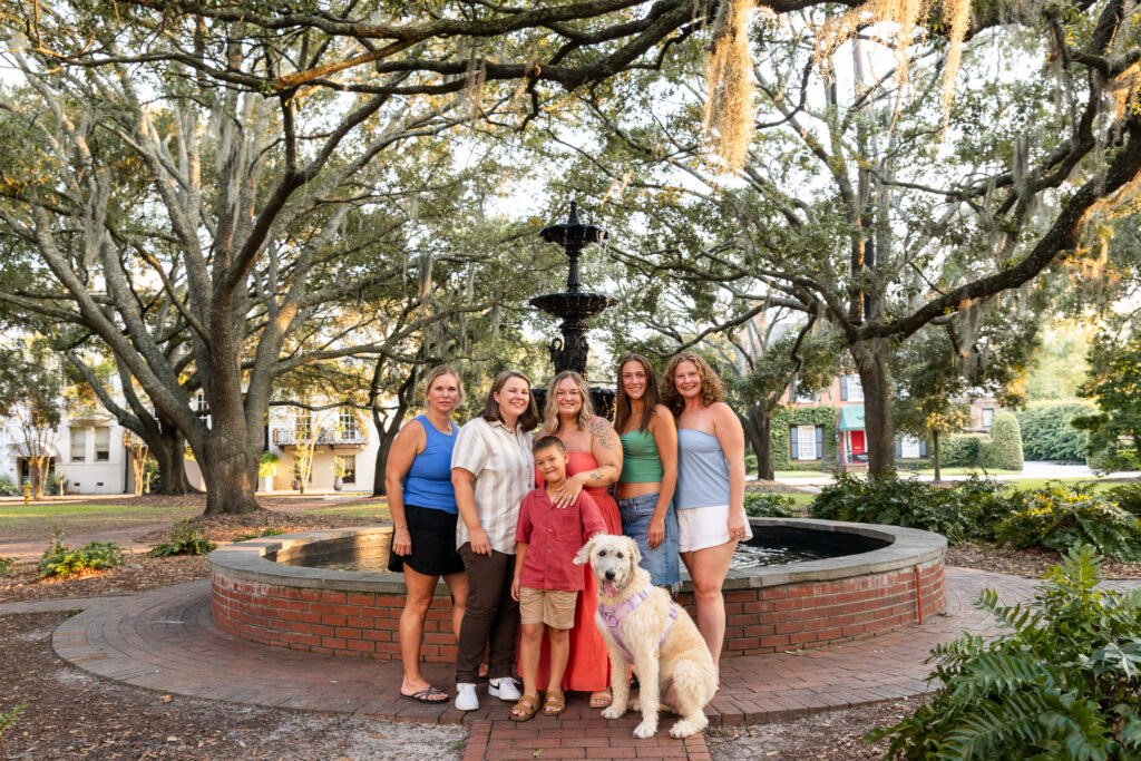 Family joins the couple to celebrate their LGBTQ proposal in Savannah, gathering for a portrait with a young boy and large fluffy white dog in front of the Lafayette Square fountain beneath moss-draped live oak trees.