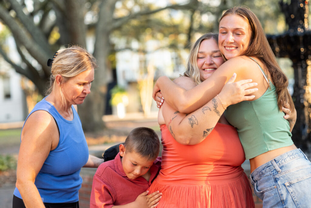 A newly engaged woman closes her eyes and smiles while tightly hugging a family member in a green top, with a young boy pressed against her side and another family member standing nearby during the celebration.