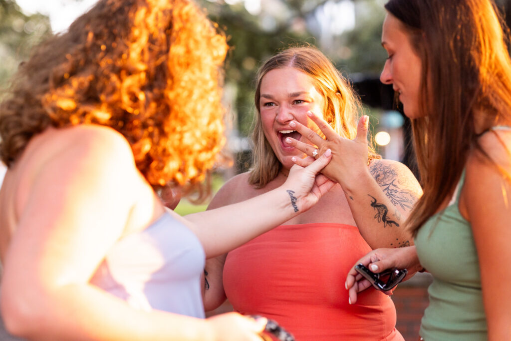 A newly engaged woman laughs as she raises her left hand between two family members, who reach in to admire the diamond ring while bright golden-hour sunlight falls across the group.