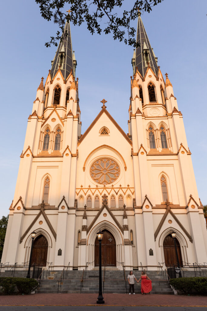 During portraits following an LGBTQ proposal in Savannah, the newly engaged couple holds hands on the sidewalk beneath the immense twin spires, pointed arches and circular rose window of the Cathedral Basilica of St. John the Baptist.